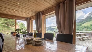 A wooden dining table with plates, glasses, and a bottle, set in a room with large windows overlooking a mountainous landscape.