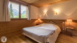 A wooden cabin bedroom with a bed, two bedside lamps, a window with curtains, and a view of mountains.