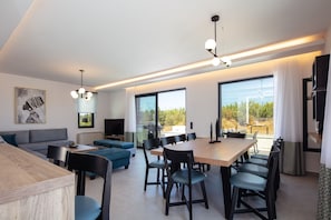 Een moderne woonkamer met een bank, een eettafel met stoelen, een televisie en grote ramen met uitzicht op groen.