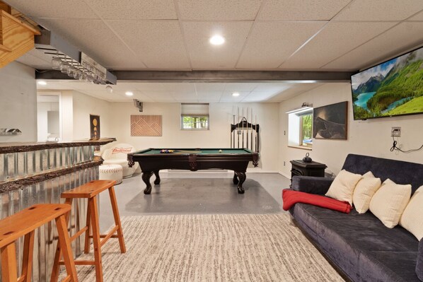 A pool table in a room with a bar area, a couch, and a television on the wall.