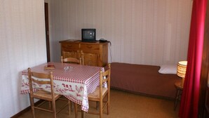 Une petite pièce avec une table et des chaises en bois, un lit, une commode avec un téléviseur et une lampe.
