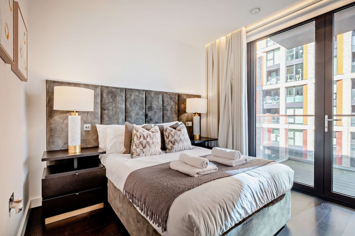 Une chambre d’hôtel moderne avec un grand lit, deux tables de chevet avec des lampes, une vue sur un immeuble à travers une porte vitrée et un balcon.
