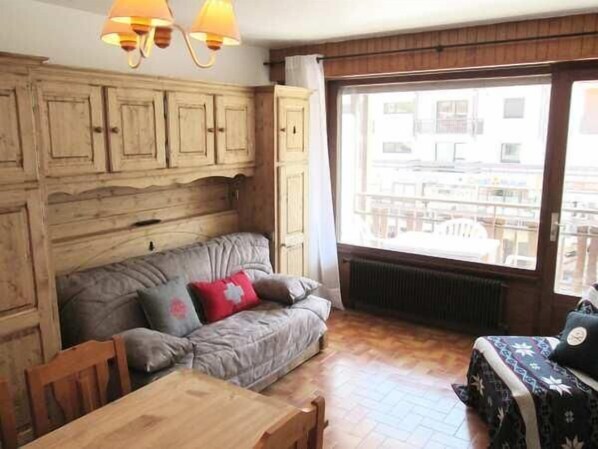 A cozy room with a wooden cabinet, a grey sofa with red cushions, a wooden table, and a large window with a view of buildings.
