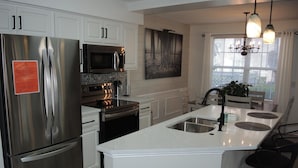 A modern kitchen with stainless steel appliances, white cabinetry, a central island with bar stools, and a large window with blinds. - 1401m 1 Bedroom Condo by Redawning (Calabash)