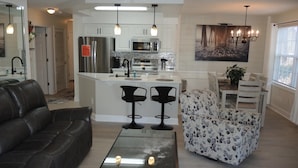 A modern kitchen with white cabinets, a central island, and stainless steel appliances. There is a dining area with a white table and chairs, and a living room with a leather couch and a glass coffee table. - 1401m 1 Bedroom Condo by Redawning (Calabash)