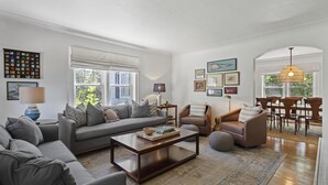 Una sala de estar moderna con un sofá gris, una mesa de centro, un comedor con mesa y sillas, y un gran ventanal con persianas.