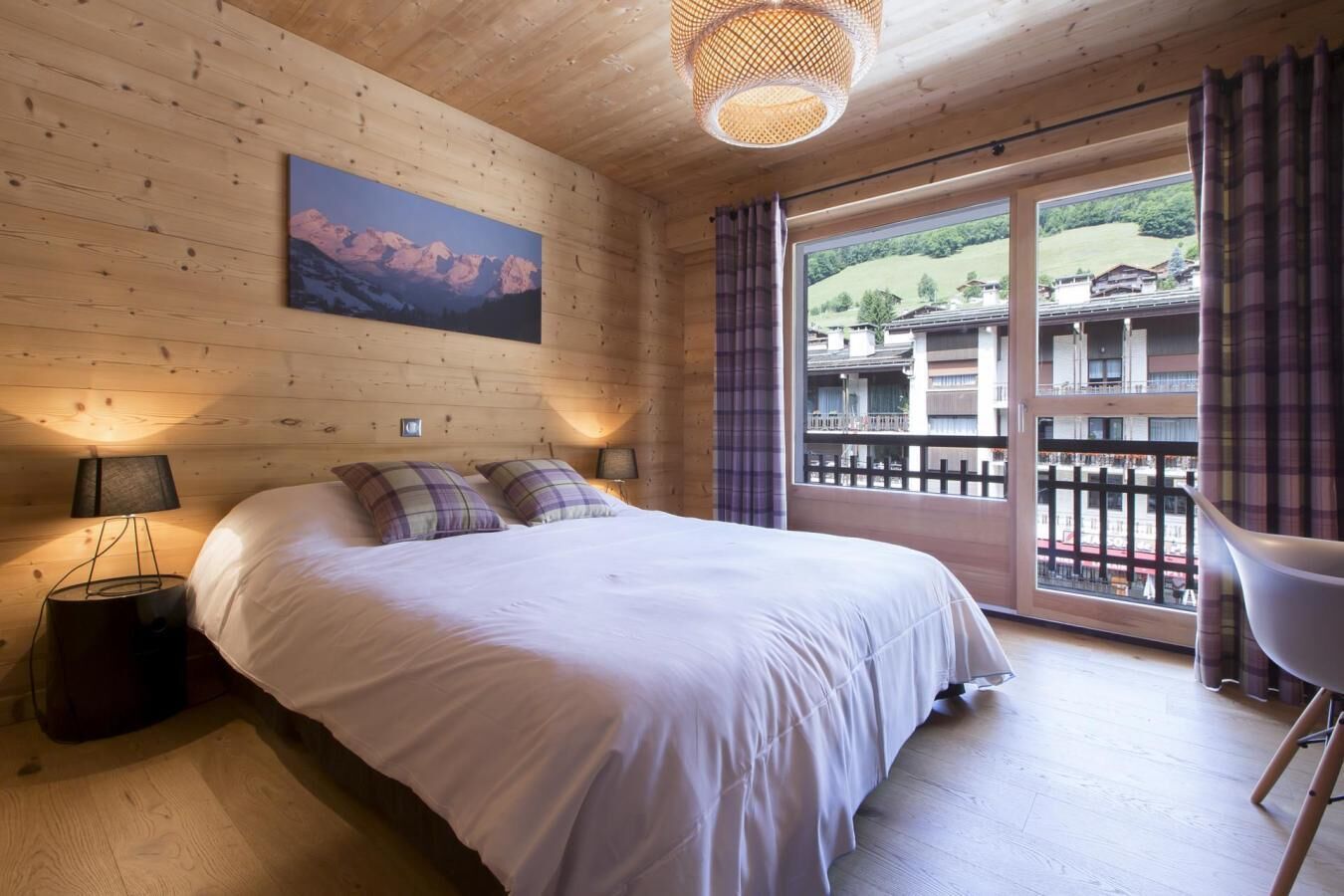 Une chambre avec un grand lit, une vue sur les immeubles depuis un balcon et un tableau accroché au mur.