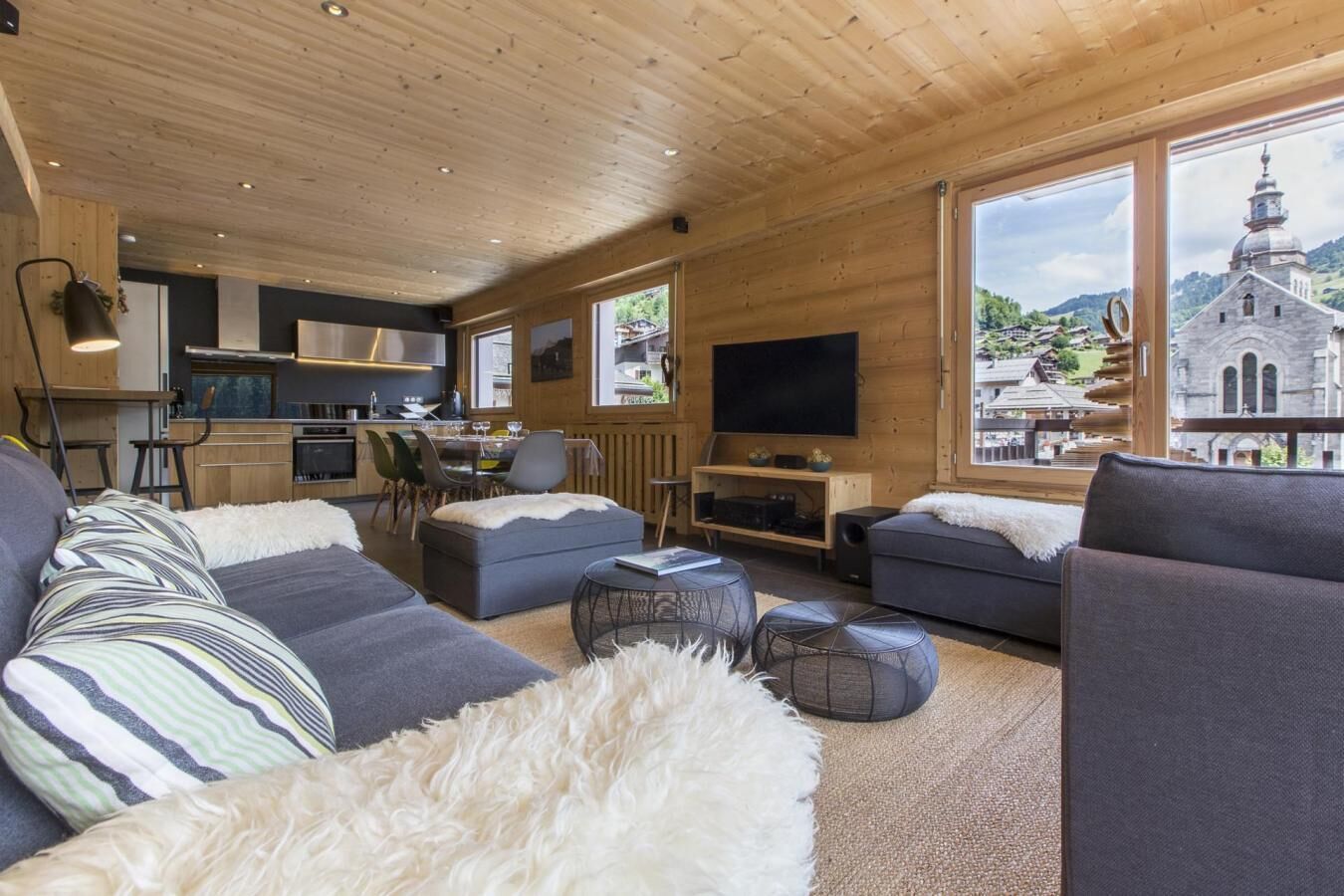 Ein modernes Wohnzimmer mit einer Couch, Hocker und einem Fernseher. Es gibt einen Essbereich mit Stühlen und einem Tisch. Große Fenster bieten Blick auf eine Kirche und Berge.