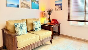 Una sala de estar con un sofá beige, tres cuadros en la pared que muestran paisajes costeros, una mesita con una lámpara y un florero, y una ventana con persianas.