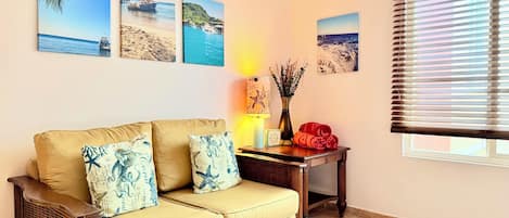 Una sala de estar con un sofá beige, tres cuadros en la pared que muestran paisajes costeros, una mesita con una lámpara y un florero, y una ventana con persianas.