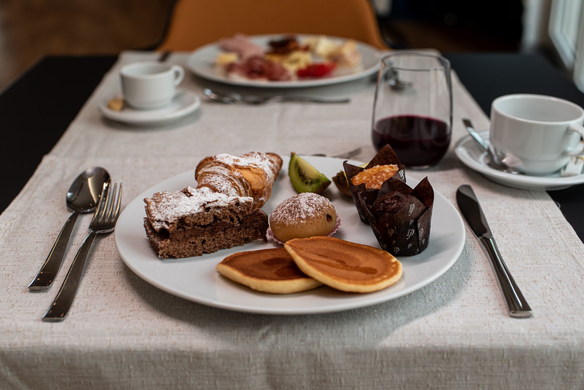 A breakfast setting with plates of pastries, a glass of red wine, and dining utensils on a table.