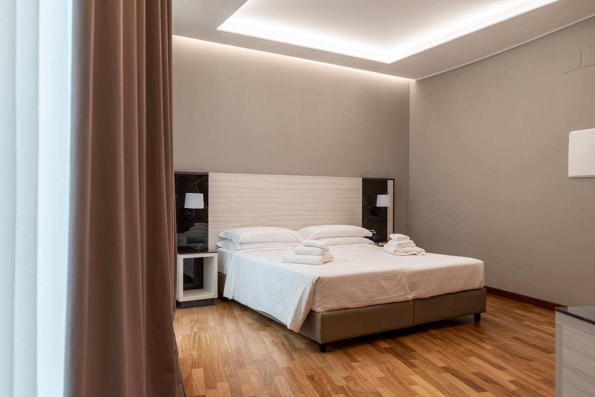 A modern hotel room with a large bed, bedside tables, a nightstand, and a ceiling light.