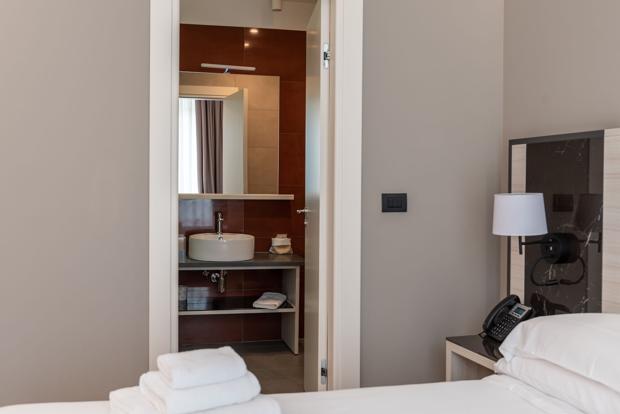 A hotel room with a bed, a bedside table with a lamp, a phone, and a bathroom visible through an open door.