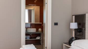 A hotel room with a bed, a bedside table with a lamp, a phone, and a bathroom visible through an open door.