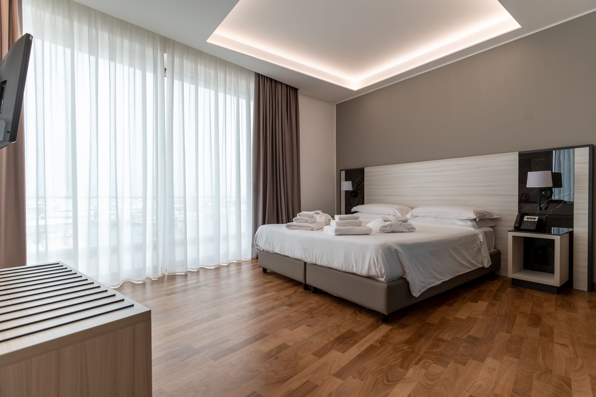Habitación de hotel moderna con una cama grande, mesitas de noche, un escritorio y vistas de la ciudad a través de cortinas translúcidas.