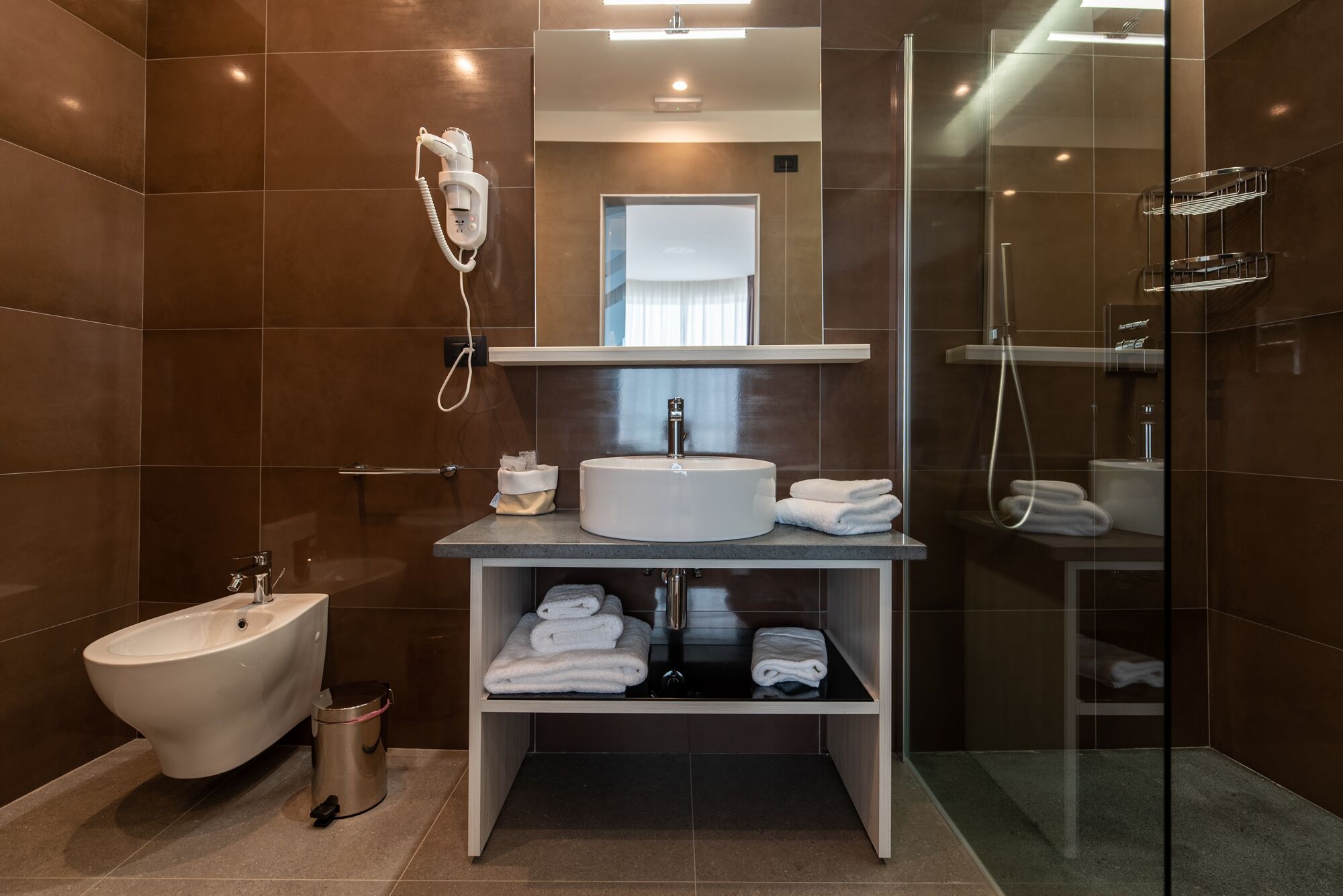 A modern bathroom with a bidet, a sink with a mirror above it, a glass shower enclosure, and a towel rack.