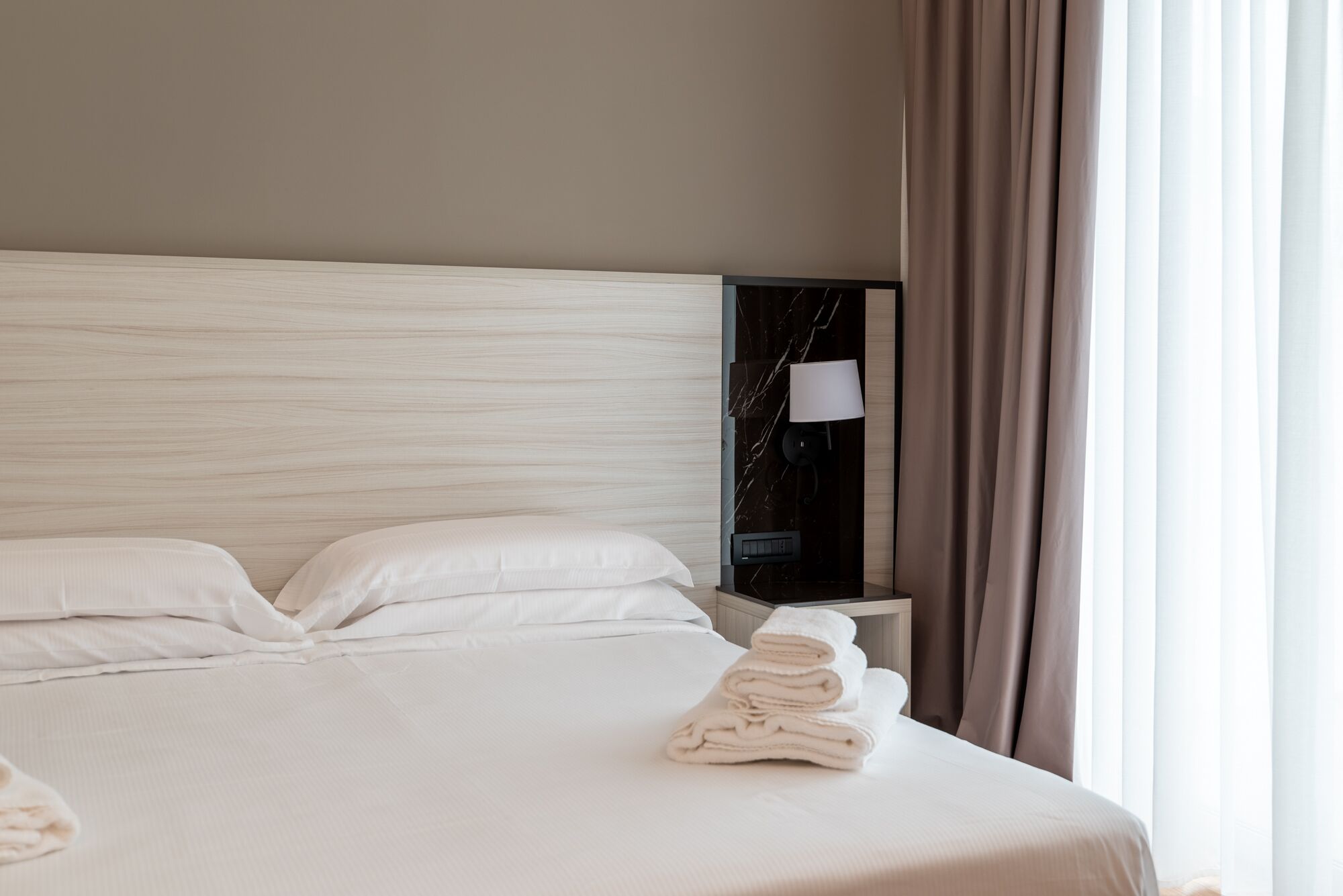 Een opgemaakt bed met witte lakens, een nachtkastje met een lamp, en uitzicht buiten door gordijnen.
