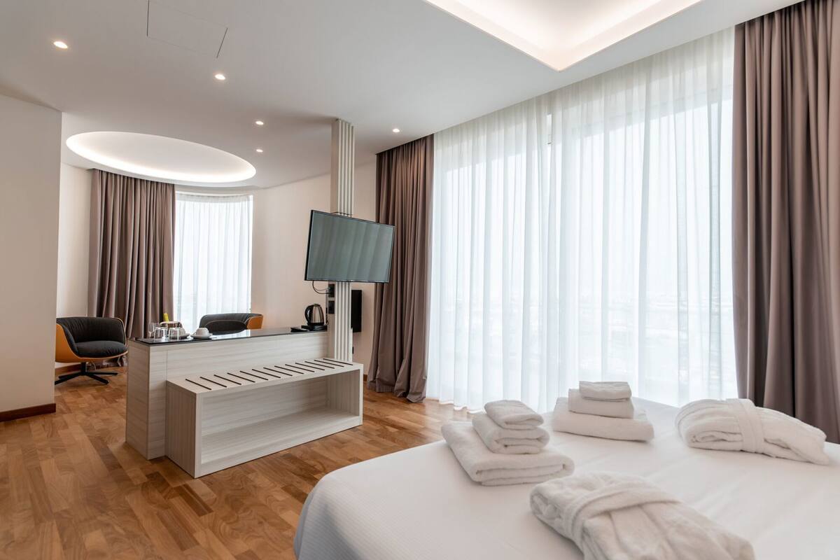 A hotel room with a bed, a desk with a TV, a chair, and a large window with curtains.