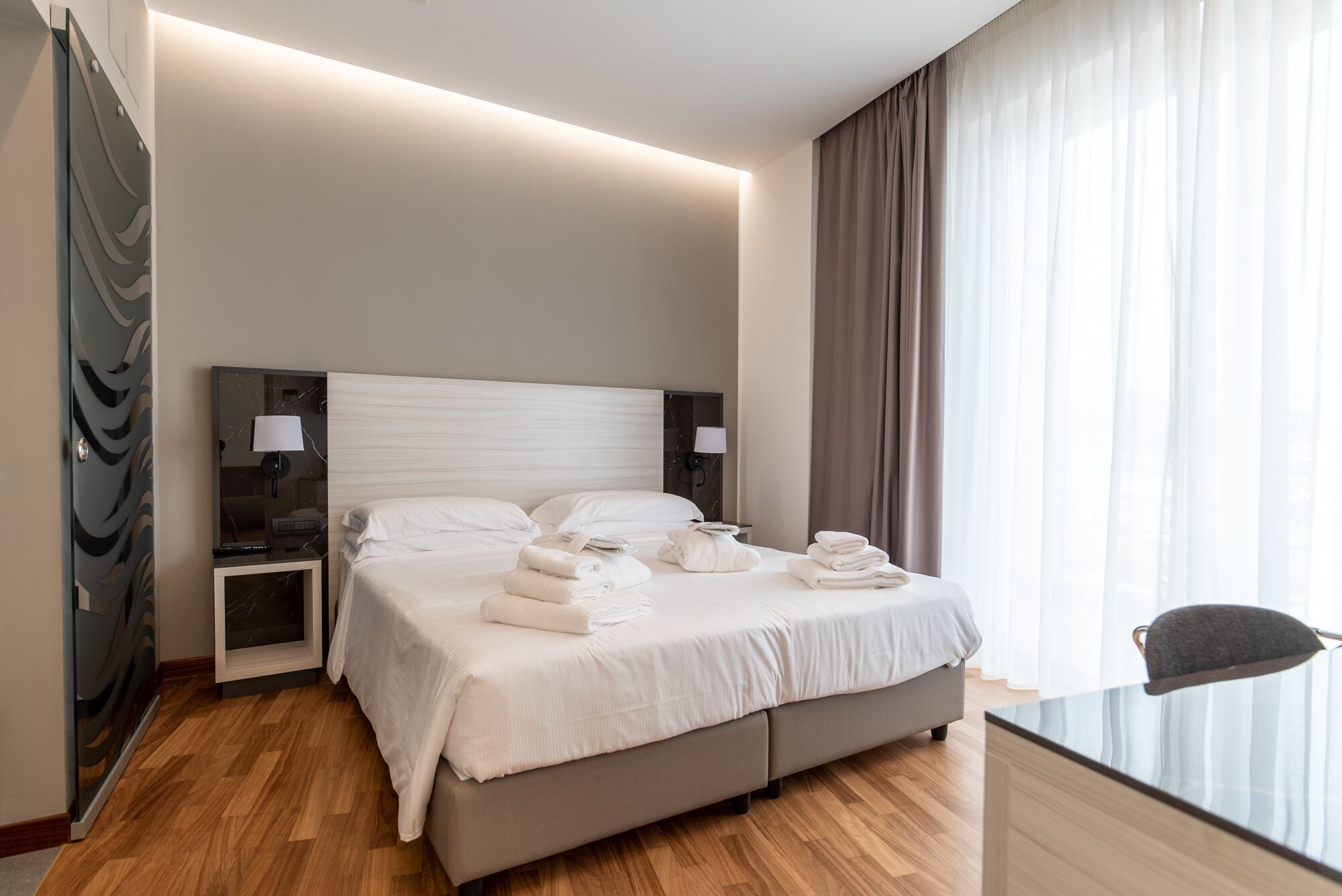 A hotel room with a large bed, bedside lamps, a flat-screen TV, and a view of the outdoors through sheer curtains.