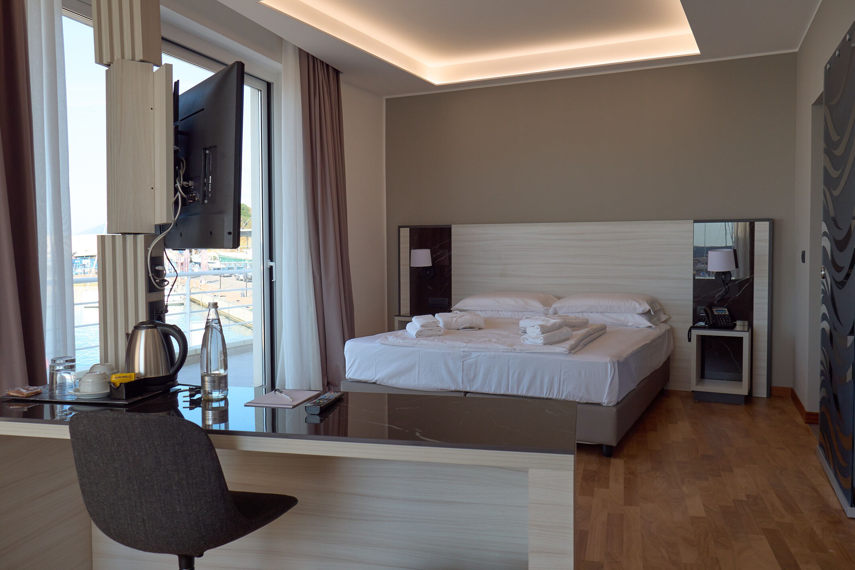 Habitación de hotel con cama, mesita de noche, escritorio, silla y un gran ventanal con cortinas.