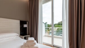 A hotel room with a bed, a nightstand with a lamp, and a view of a balcony with greenery.