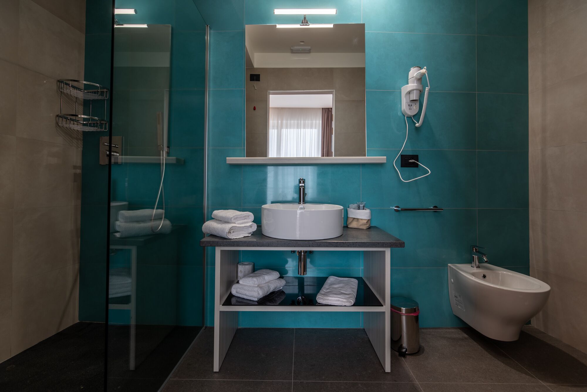 A modern bathroom with teal tiles, a glass shower enclosure, a white sink with a mirror above, a bidet, and a towel rack.