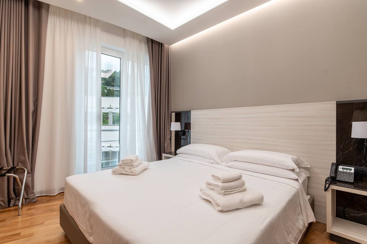 A modern hotel room with a large bed, a bedside table with a phone, and a view of greenery outside.