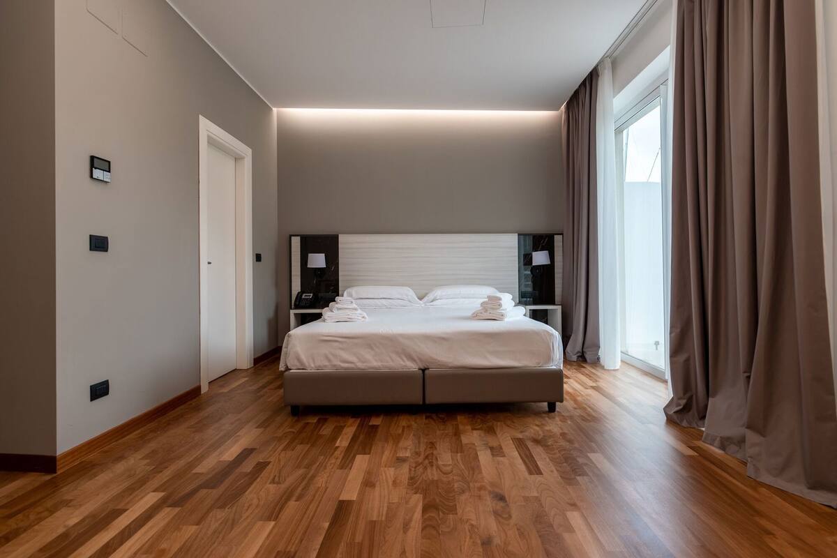 A modern hotel room with a large bed, bedside lamps, a control panel, and a view of the outdoors.