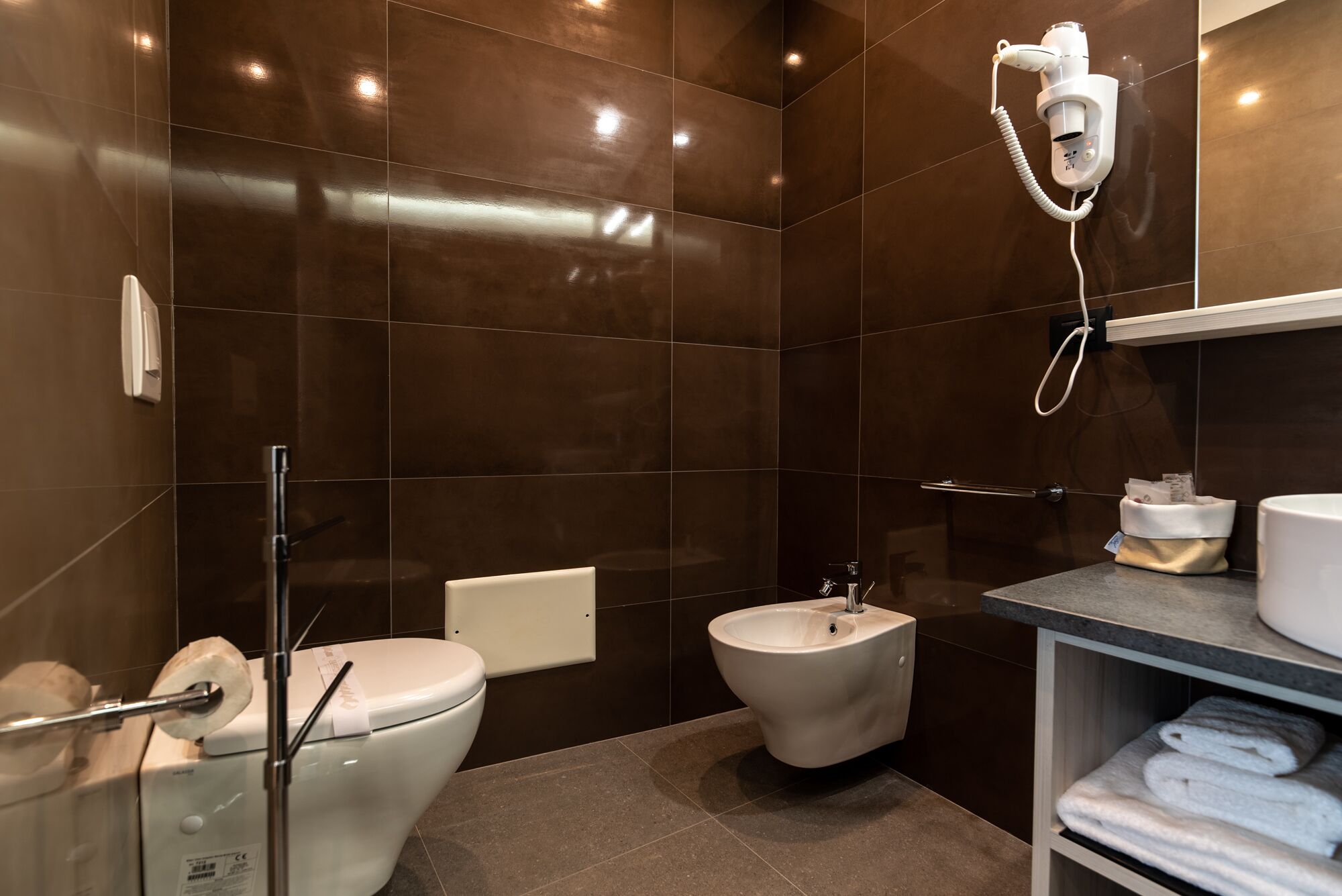 A modern bathroom with dark tiles, a toilet, bidet, and a towel rack.