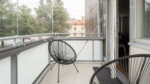 Un balcón con dos sillones negros de mimbre, barandilla de vidrio y vista a edificios y árboles al fondo.
