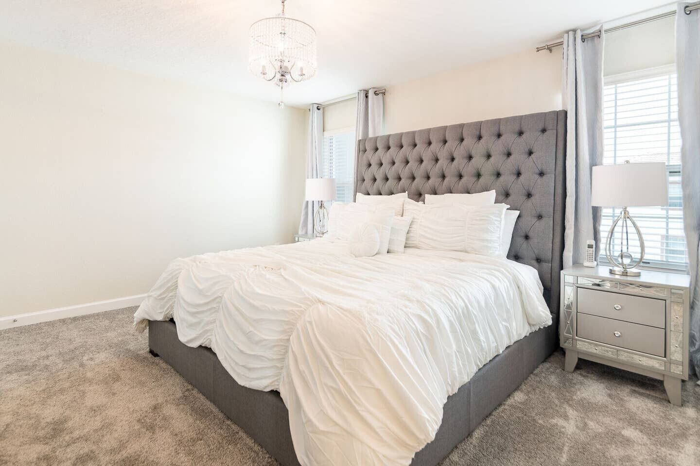 Ein Schlafzimmer mit einem großen Bett, einem gepolsterten Kopfteil, einer Kandelaberleuchte, einem Nachttisch mit Lampe und einem Fenster mit Vorhängen.