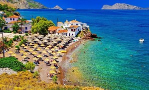 A coastal village with white buildings, clear blue waters, and a beach with umbrellas.