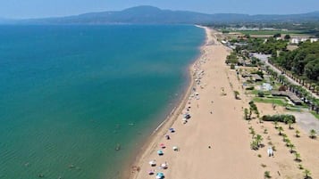 Eine Luftaufnahme eines Strandes mit einer weiten Sandfläche, vereinzelten Sonnenschirmen und einer Baumreihe entlang der Küste.