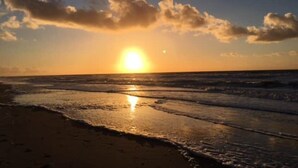 Une plage au coucher du soleil, avec le soleil bas à l’horizon, des nuages dans le ciel et des vagues qui se rapprochent du rivage.