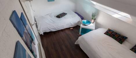 A small, compact room with two beds, a small table with a bowl and a plant, and a painting on the wall.