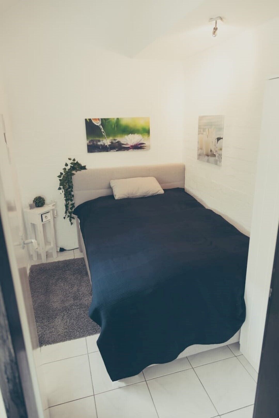 Een kleine, verzorgde slaapkamer met een bed, een klein nachtkastje en een aan de muur hangend schilderij.
