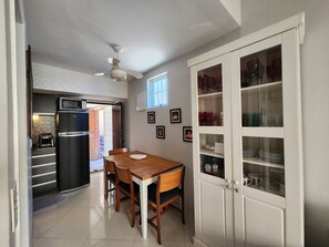 A compact kitchen with a refrigerator, a dining table with chairs, a ceiling fan, and a cabinet with glass doors. - AP Pepê Jacuzzi/solário (Rio de Janeiro)