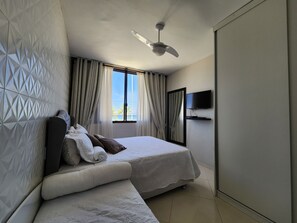 Une chambre d’hôtel moderne équipée d’un lit, d’un ventilateur de plafond, d’un téléviseur fixé au mur et d’une fenêtre avec des rideaux.