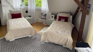 Quarto com duas camas, uma janela com cortinas, uma cadeira, mesa de cabeceira e apoio para objetos pessoais.