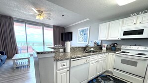 A modern kitchen with white cabinets, a granite countertop, and a large island. There is a ceiling fan, a flat-screen TV mounted above the island, and a view of the ocean through large windows. - Beach Club D0302- Direct Gulf View - Beach - Pool - WIFI (Gulf Shores)