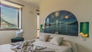 A bedroom with a large bed, a window with a mountain view, a painting of a sunset over water, and a green vase with flowers. - Varenna La Bomboniera (Varenna)