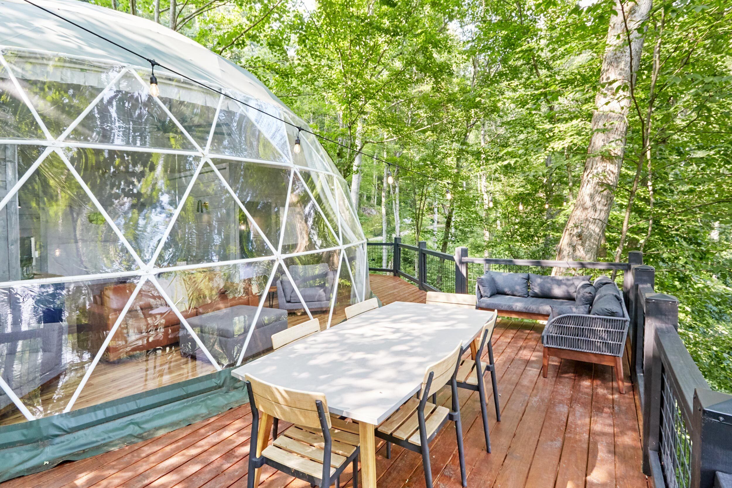 A geodesic dome structure with a wooden deck, outdoor seating, and a view of a forested area.