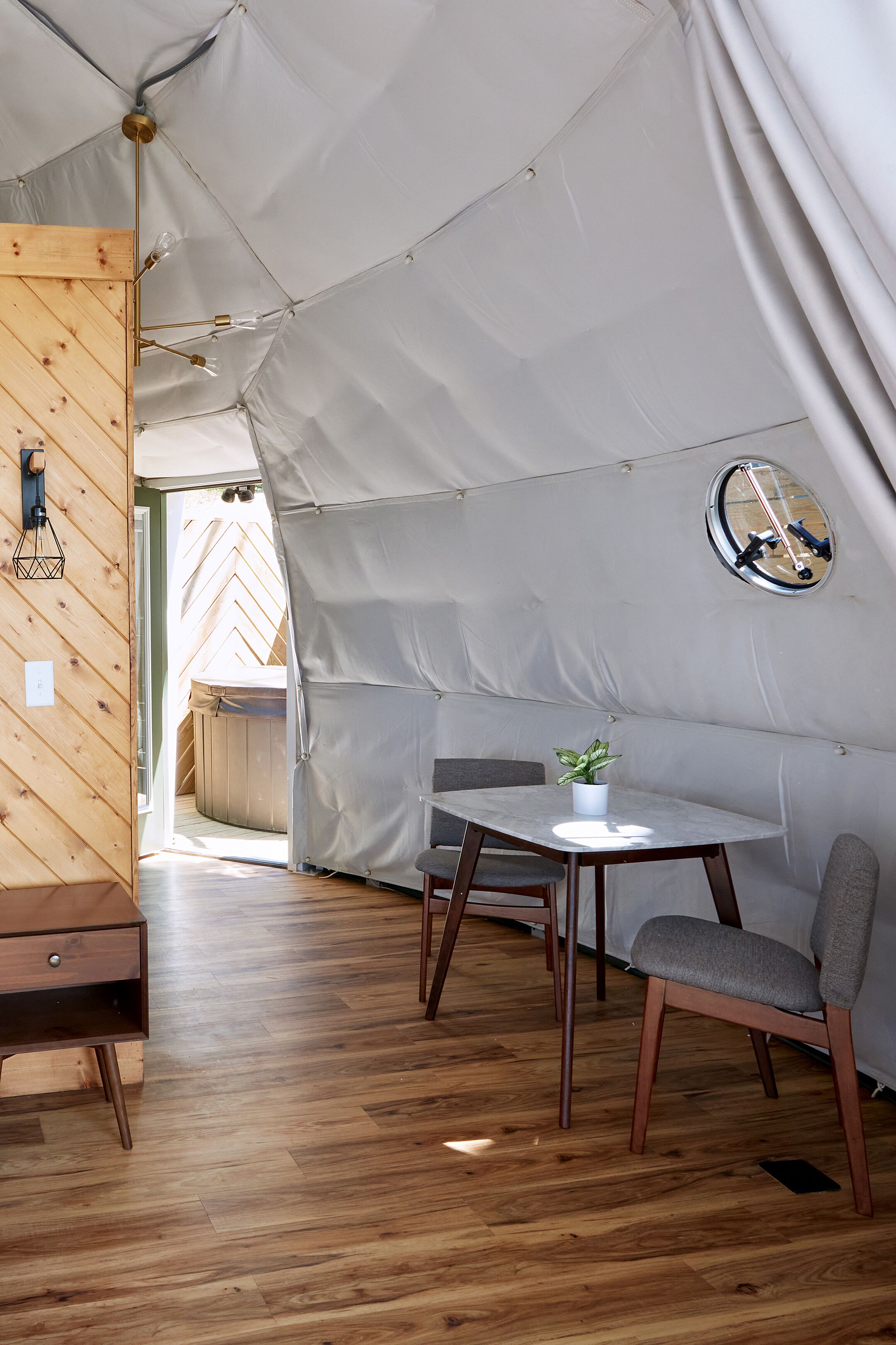 Una habitación con carpa, suelo de madera, una mesa con planta en maceta y un espejo redondo en la pared.