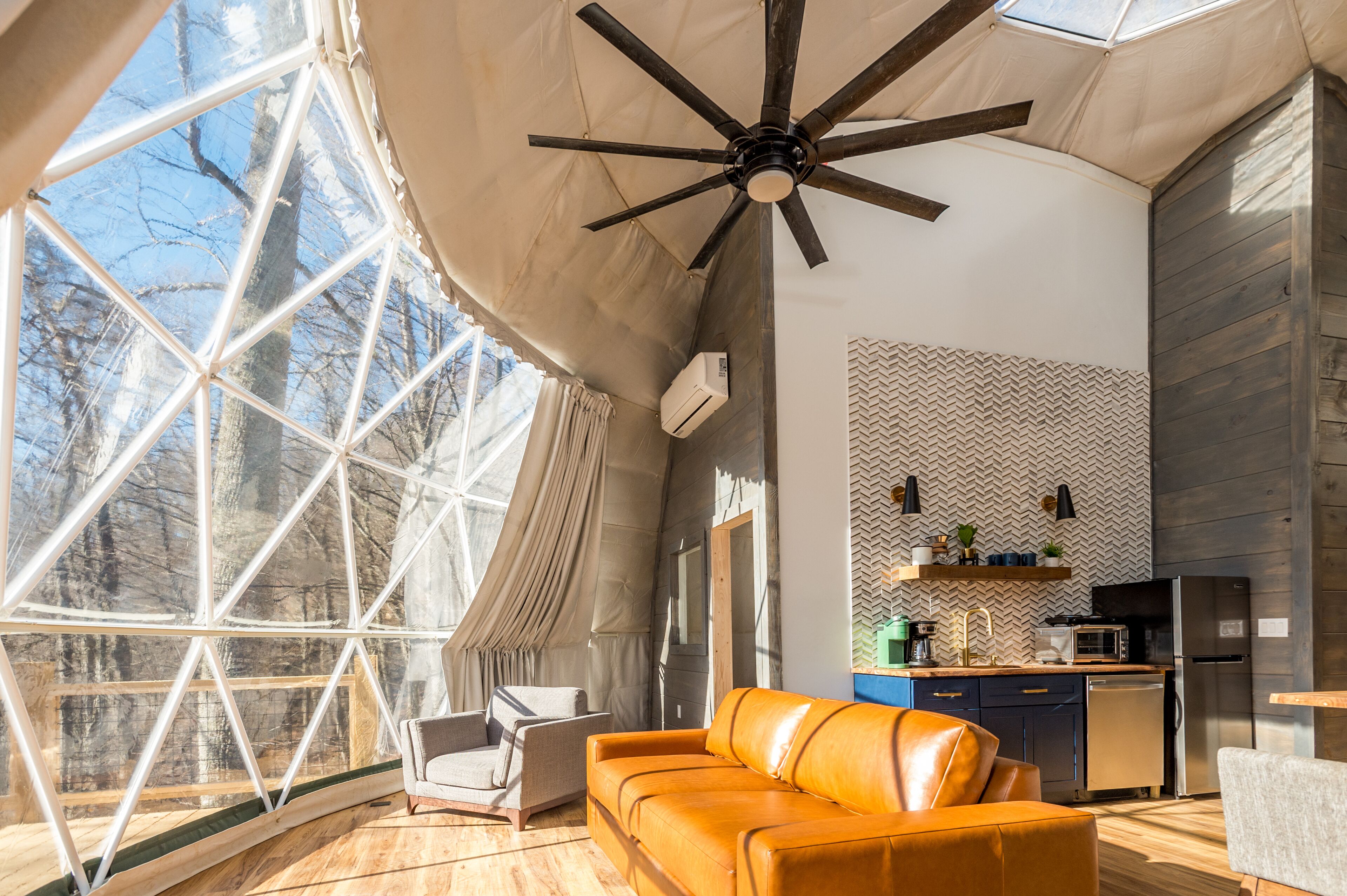 A spacious tent with a ceiling fan, a sofa, a chair, a kitchen area, and large windows offering a view of trees.