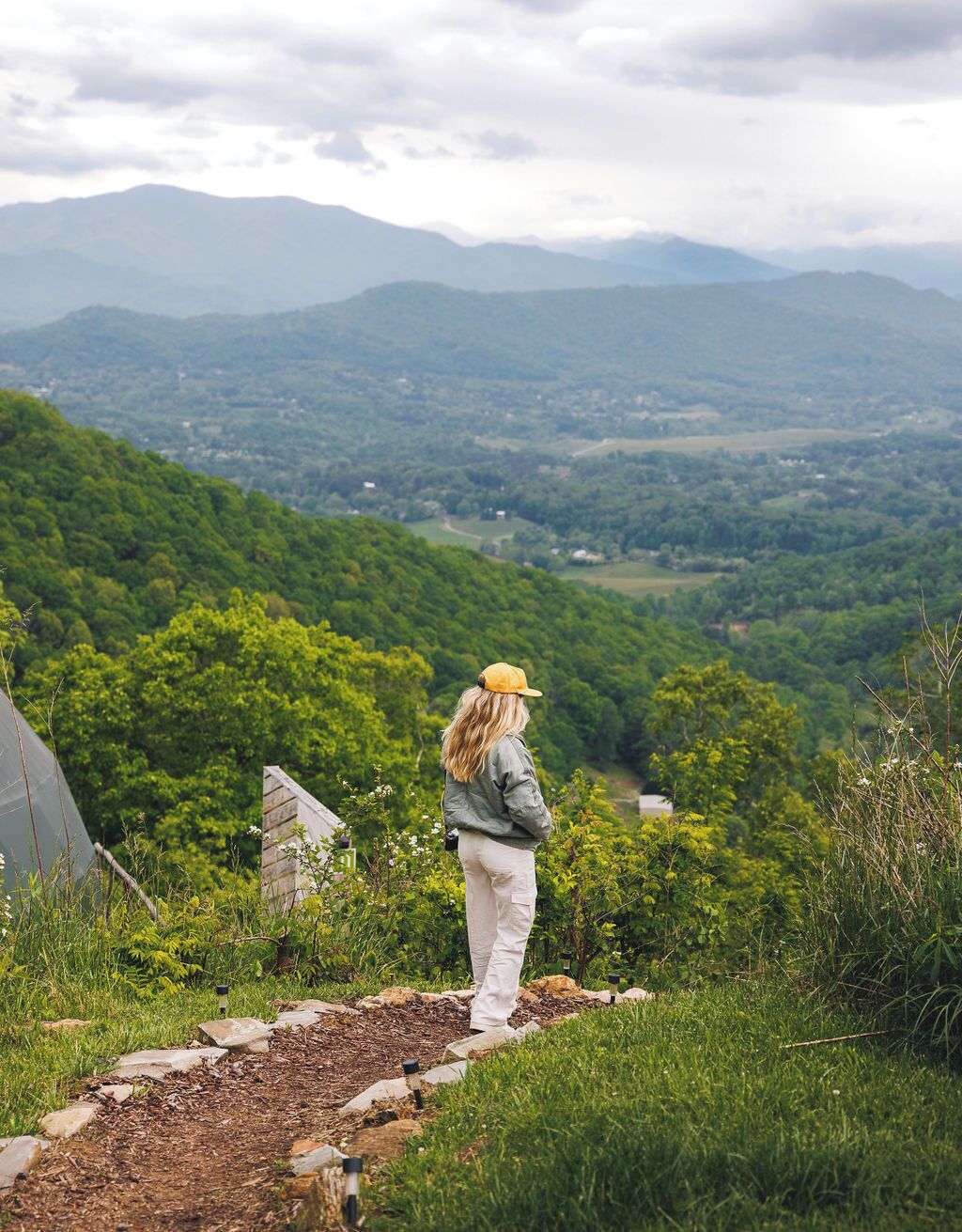A person standing on a stone path overlooking a lush valley with distant mountains.
