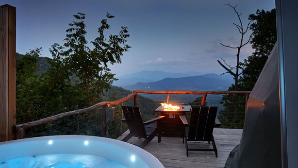 Jacuzzi sobre un pont de fusta amb una taula de foc i cadires, amb vista a un paisatge de muntanya.