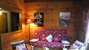 A cozy room with a wooden interior, a red sofa with patterned cushions, two wooden chairs, a round wooden table with a floral centerpiece, and a wall-mounted lamp.