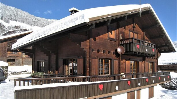 A wooden chalet with a balcony decorated with heart-shaped ornaments, set against a snowy mountain backdrop.