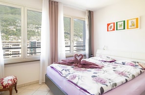 Ein Hotelzimmer mit Bett, Sessel, Fenster mit Vorhängen und Blick auf eine Berglandschaft.