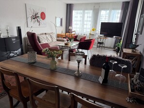 Un salon moderne avec une table à manger en bois, des chaises rouges à motifs, un canapé, un téléviseur et un oiseau décoratif posé sur une table d’appoint.